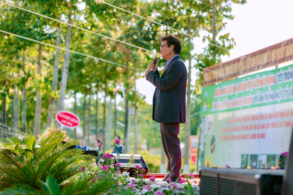 A man in a dark suit holding a microphone stands on an outdoor stage surrounded by lush green trees and potted plants with pink, purple, and white flowers. The stage backdrop displays a large, colorfu