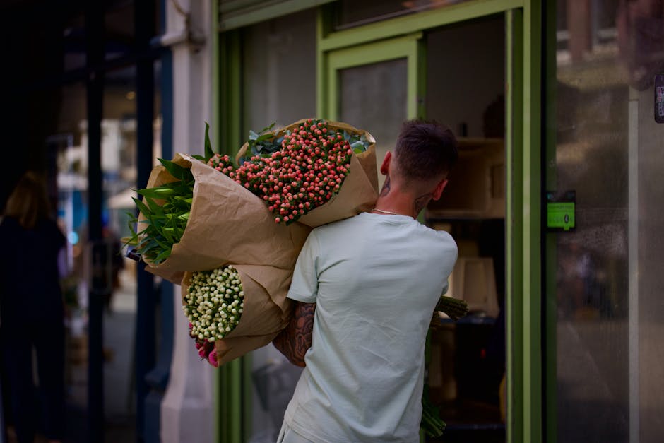 A person wearing a light grey t-shirt holds a large bouquet of fresh flowers outside a flower shop with green-framed glass doors. The bouquet includes pink and white waxflowers, with lush green foliag