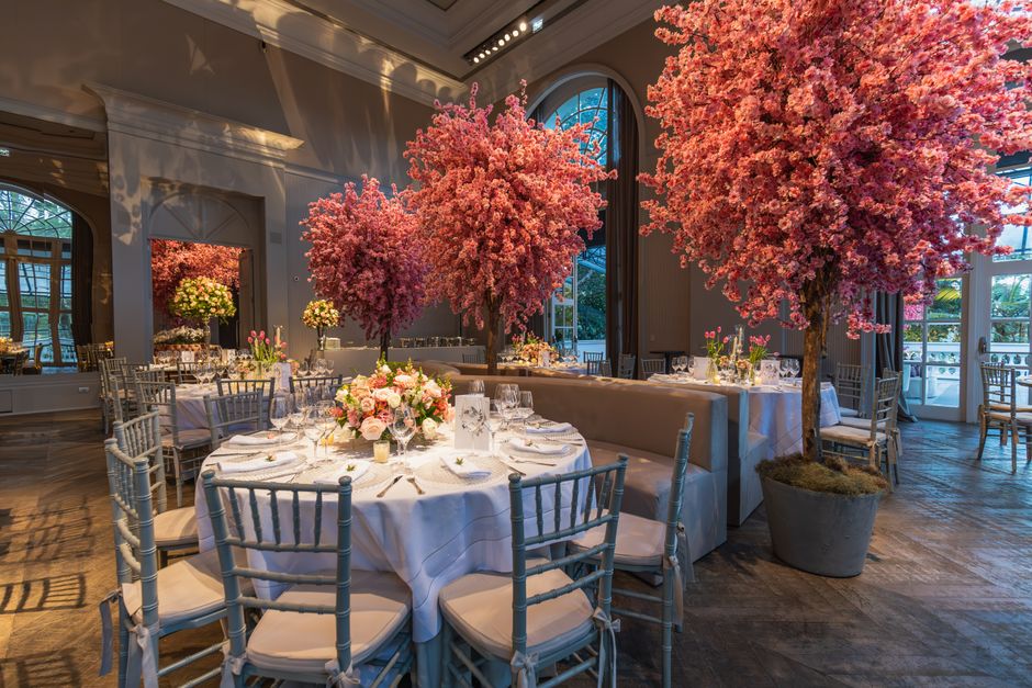 The image showcases an elegant event space with large, vibrant pink cherry blossom trees in full bloom, placed in decorative pots around the room. The area is illuminated by natural light streaming th