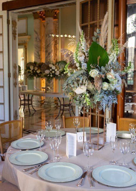 A formal wedding reception table set with a large, elegant floral arrangement featuring white roses, pale green chrysanthemums, and pampas grass, accented with large green leaves and muted silver foli