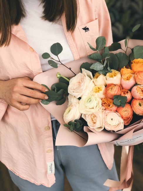 A person holding a large bouquet of fresh flowers wrapped in pale pink paper, featuring white and peach ranunculus, soft pink tulips, and green eucalyptus leaves. The person is wearing a light pink sh